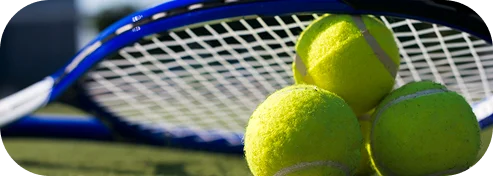 Une raquette de tennis avec un cadre bleu est posée sur une surface avec trois balles de tennis jaunes. Le court de tennis est ensoleillé avec une ligne blanche en bas.