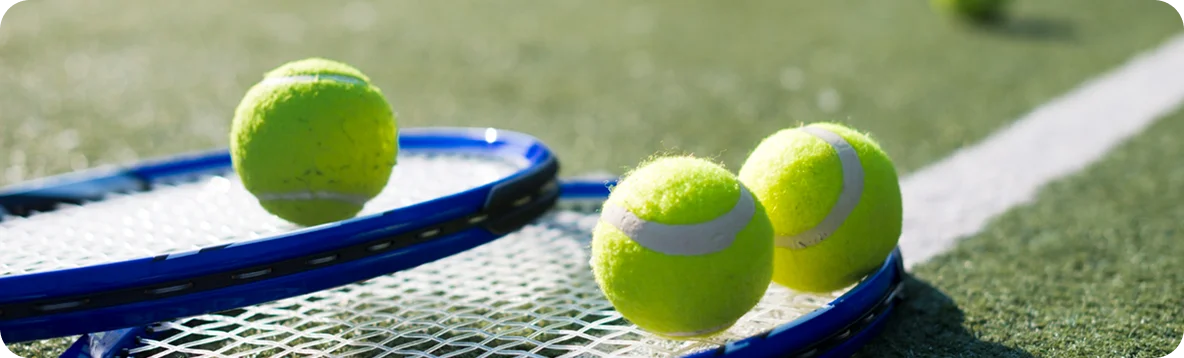 Trois balles de tennis jaune vif reposent sur et autour d'une raquette de tennis au cadre bleu. La scène se déroule sur un court de tennis ensoleillé où l'on aperçoit une ligne blanche.